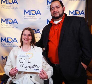 Amy and her partner Jon pose in front of an MDA backdrop while Amy holds a sign that says "I advocate for GNE myopathy."