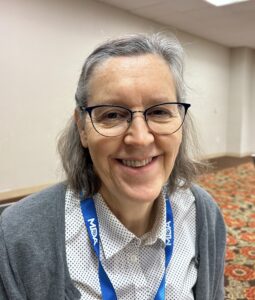 Standing female smiles with grey hair, glasses, blue neck lanyard, white shirt and grey cardigan