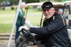 Mike Bellotti in smiles from a golf cart wearing a black ball cap and black long sleeve athletic shirt