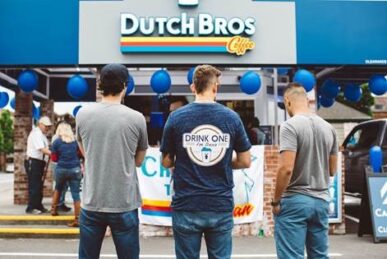 Backview of three men standing in front of a Dutch Bros location, one man wearing a Drink One for Dane T-shirt