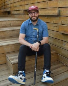 Billy Zureikat, a white man with tortoise shell-rimmed glasses and a brown beard, sits on the stairs of a wooden deck, wearing a red baseball cap, blue denim button-down shirt, black jeans, and blue and white high-top Nike sneakers, and holding a walking cane.