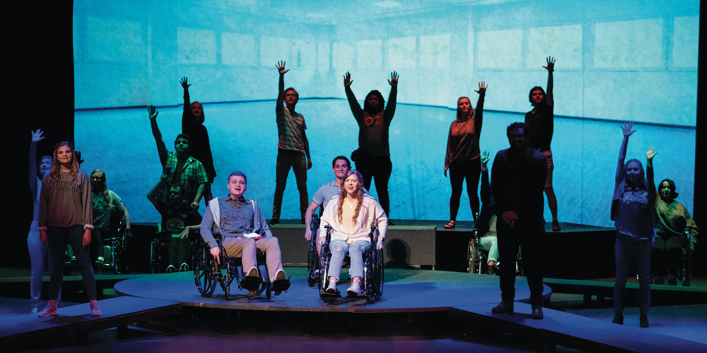 A scene in “Wheels: An Original Musical,” where 17 people can be seen on stage facing the audience, some sitting in wheelchairs and some standing with a hand raised in the air against a blue backdrop.