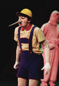 Jess Westman, in costume as Pinocchio, wears a yellow cap, long wooden nose strapped around his cheeks, yellow shirt with blue overalls, sleeves with faux wood grain, and white gloves. 