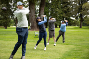 A man, woman, and two other men stand in a line swinging golf clubs outside.