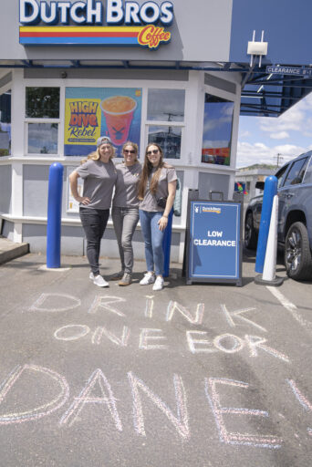 Katie Hutchinson, a Dutch Bros teammate, and Kinsey Fischer stand in front of a Dutch Bros location with "Drink One for Dane" written in chalk on the parking lot