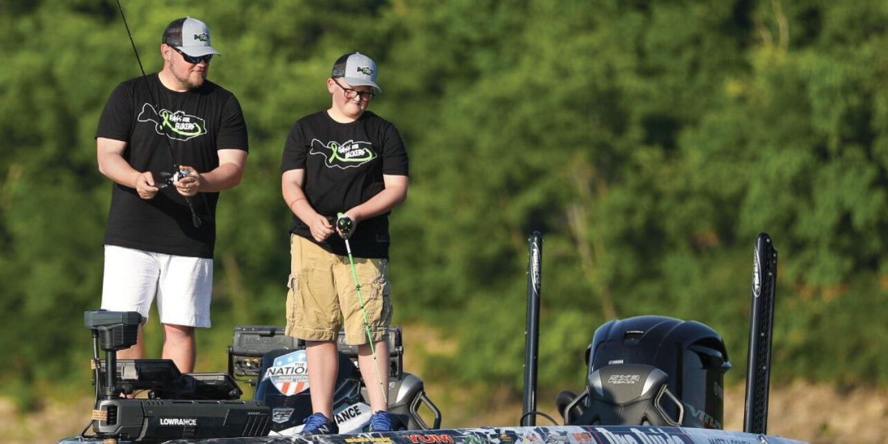 Matt Looney and his teenage nephew Talon stand on the deck of a fishing boat, each wearing black Bass for Beckers T-shirts and matching grey caps.