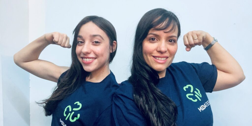 Leah and her mother, Bevi, pose back to back each holding a flexed arm in the air