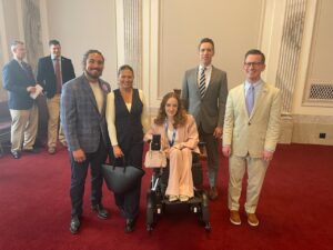 A young woman in a wheelchair with a man and woman standing on one side and two men, one a Senator, standing on her other side