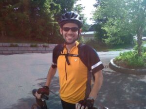 Nevin Steiner smiles wearing a bike helmet stands with his bike and road behind him