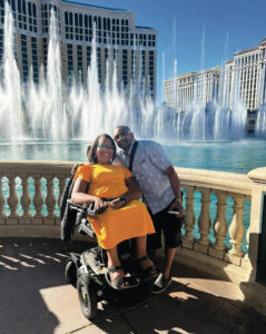 Leticia and her husband pose in front of the Fountains of Bellagio in Las Vegas. She is a Black woman with shoulder-length dark brown hair sitting in a power wheelchair, wearing a bright yellow short-sleeved dress and sandals. He is a Black man with close-trimmed black hair and beard, wearing a blue button-down short-sleeved shirt and black shoes.