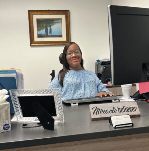 Letitia Tatum, a Black woman with long black hair wearing green glasses and a light blue shirt, sits at an office desk, with her left hand on a keyboard. A sign on the desk says “Miracle believer.”