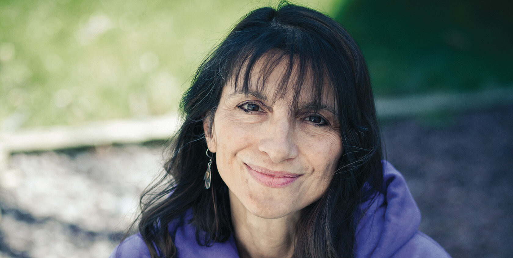 Closeup of Jodi O’Donnell-Ames, a white woman with long black hair, sitting with her arms around her knees, wearing a purple Hope Loves Company sweatshirt.