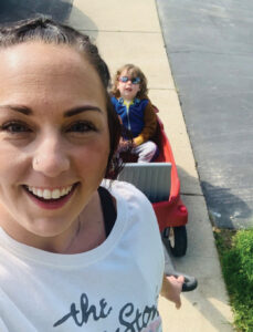 Closeup of Sabrina Johnson, a white woman with dark brown hair pulled back. Just behind her, her son, a boy with shoulder-length light brown hair and sunglasses, sits in a red wagon.
