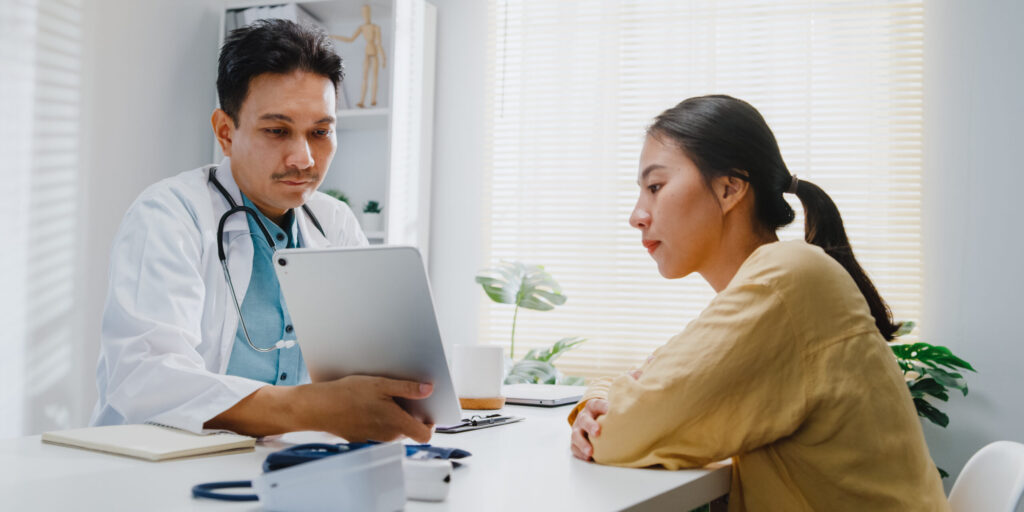 Mature Asian man doctors medical results during consultation on tablet computer with woman patient in health clinic. Medical health care concept.