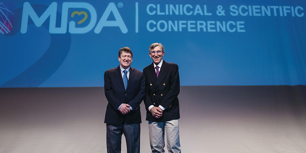 Two men in dark suit jackets and ties stand in front of a large blue screen with words projected on it: MDA Clinical & Scientific Conference.
