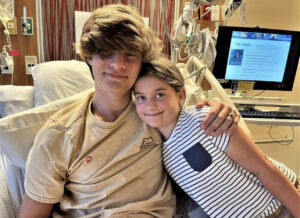 A young girl in a striped shirt smiles next to a teenage boy in a hospital bed
