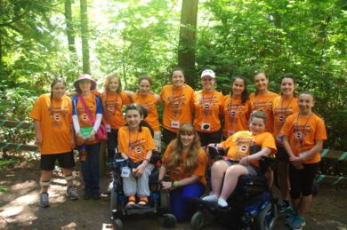 A group of individuals standing and sitting in wheelchairs wear matching orange MDA Camp t-shirts