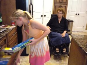 In Barbara Twardowski’s kitchen, a teenage girl opens a drawer to get a kitchen tool while the author observes, sitting in her power wheelchair.
