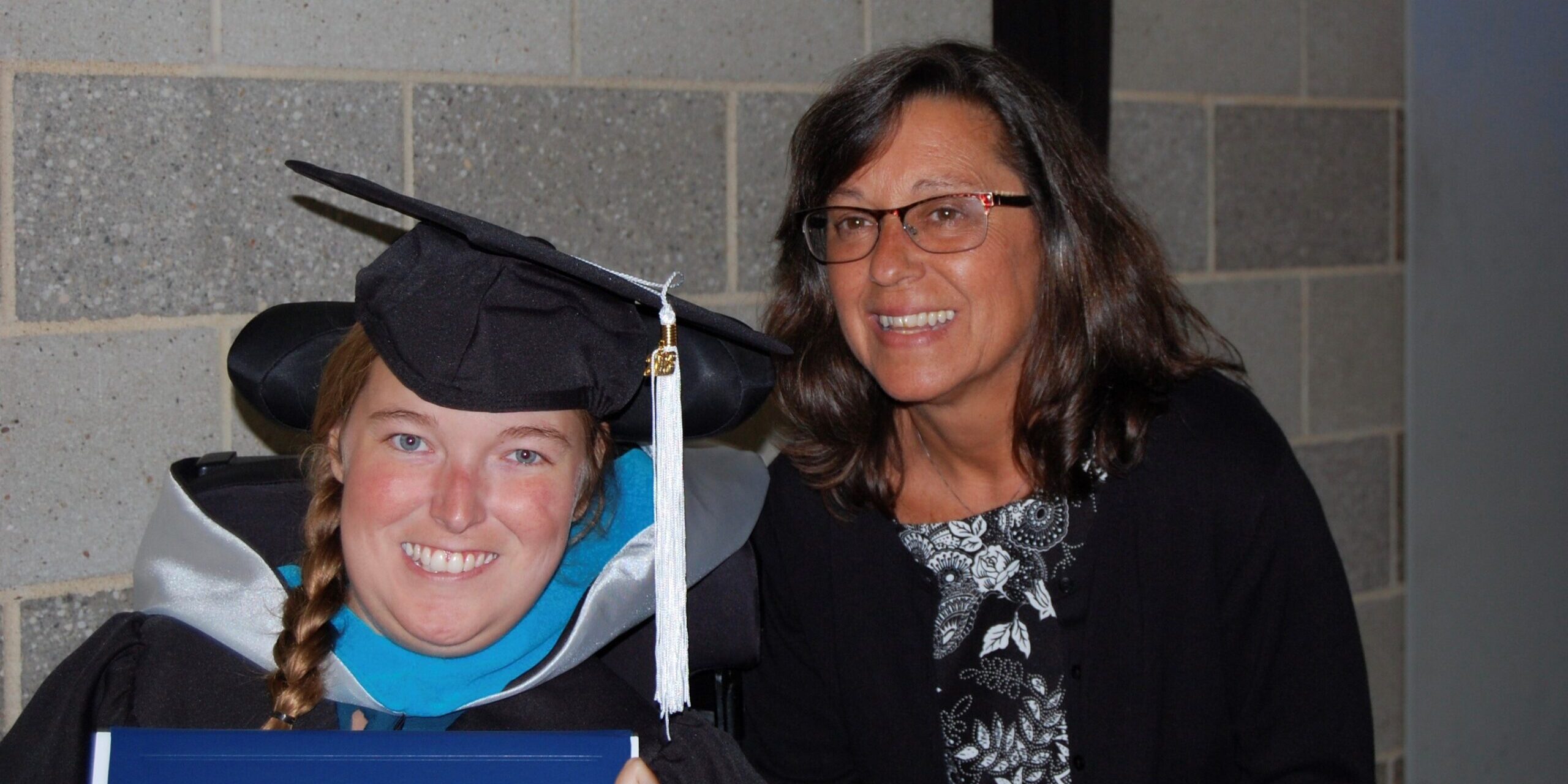 A young woman in a wheelchair wears a graduation cap and gown while holding a diploma and her mother smiles next to her