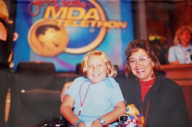 A young girl in a wheelchair and her mother smile in front of the MDA Telethn backdrop