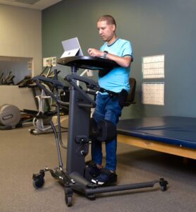 Jose Flores is in an upright position within a stander that uses straps and padding to support the legs and torso, while he rests his arms on a tray at chest height. Wheels on the base of the stander make it mobile.