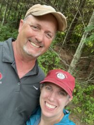 A husband and wife smile wearing baseball hats outside