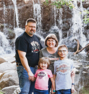 Family of four smiles in matching Harley Davidson t-shirts in front of waterfall holding hands. 