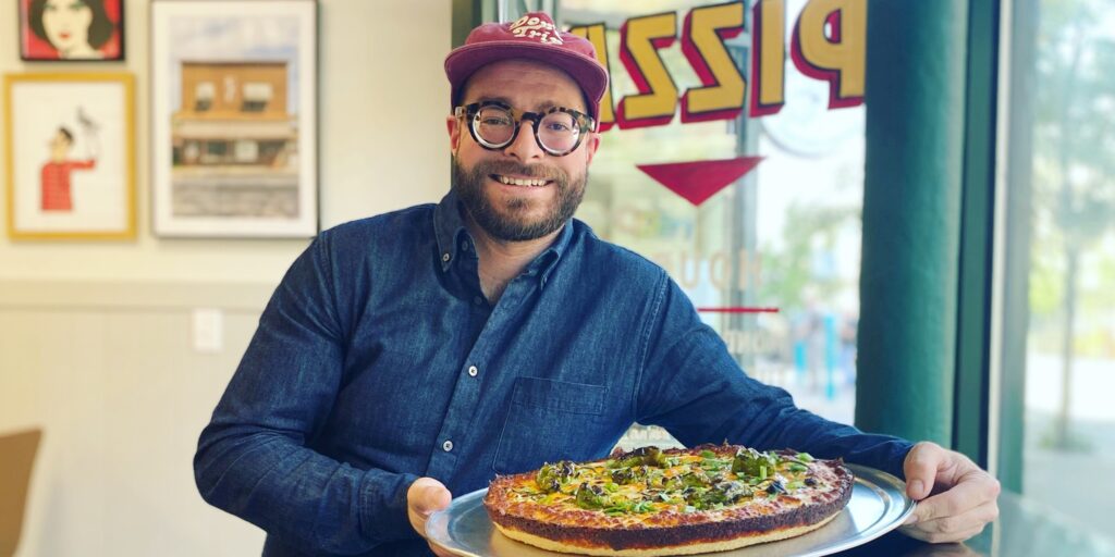 WHITE MAN WITH GLASSES, RED HAT, AND BLUE SHIRT HOLDING A PIZZA