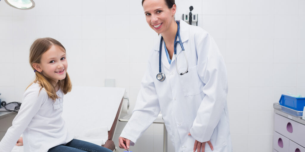 A young girl with blonde hair sitting on a doctor’s table as a doctor, a woman with brown hair, uses a reflex hammer on the girl’s knee. Both girl and doctor are smiling.