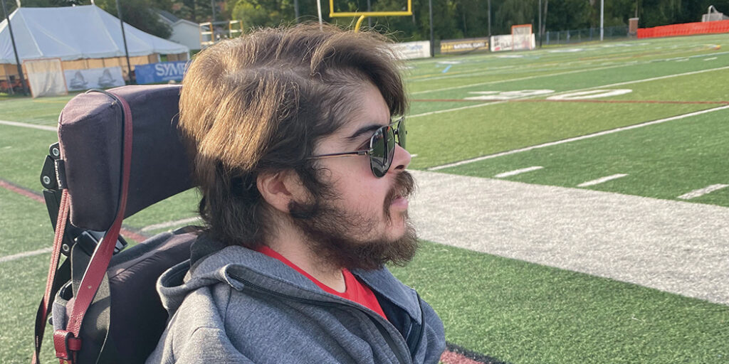 Jonathan Piacentino, with brown hair and a beard wearing a grey zip-up sweatshirt with a red shirt underneath, sits in his wheelchair looking into the distance on a football field.