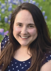 Closeup photo of Brooke Aarvig with brown hair and a blue printed blouse in front of a field of flowers. 
