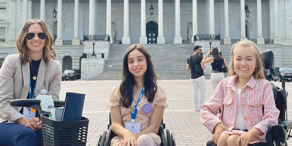 Amy Shinneman wearing a beige blazer, Leah Zelaya wearing an orange floral dress and Reagan Imhoff, in a pink and white tweed blazer, sit smiling in their wheelchairs with their hands in their laps in front of the US Capitol building.