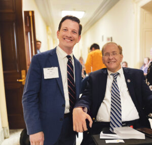 Michael Lewis, a white man with brown hair, smiles while standing next to Rep. Jim Lang, who is seated in a wheelchair and also smiling. Both men are wearing suits with striped ties. 