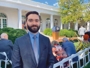 Paul Melmeyer, a white man with brown hair and a beard, smiles while standing with the White House Rose Garden behind him. He is wearing a dark suit and tie. The Rose Garden is set up for a press conference with a podium and chairs, some with people sitting in them. 
