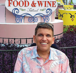 An image of Ken Yorgan, a 59-year-old man with blue eyes and salt-and-pepper hair. We see Ken from the shoulders up, smiling and wearing a pink Hawaiian-print shirt. Behind him is a sign for a Food and Wine Festival.