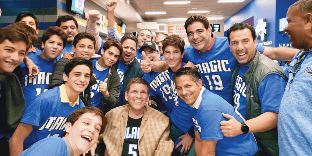 A photo of Ken Yorgan surrounded by a group of men and boys wearing blue Orlando Magic shirts. Everyone is smiling. Yorgan is seated in the middle, wearing a black Orlando jersey under a tan plaid blazer. Ken is a 59-year-old man with blue eyes and salt-and-pepper hair.