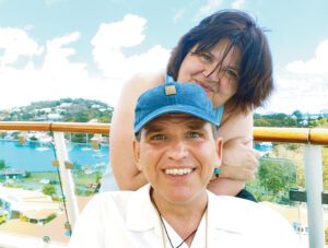 A photo of Ken Yorgan sitting with his wife, Susan, leaning behind him. Both are smiling. Ken is a 59-year-old man with blue eyes and salt-and-pepper hair, and he is wearing a white shirt and a blue-baseball hat. Susan has short brown hair and blue eyes. She is wearing a tank top. Behind them, you can see a tropical setting with water and boats. 