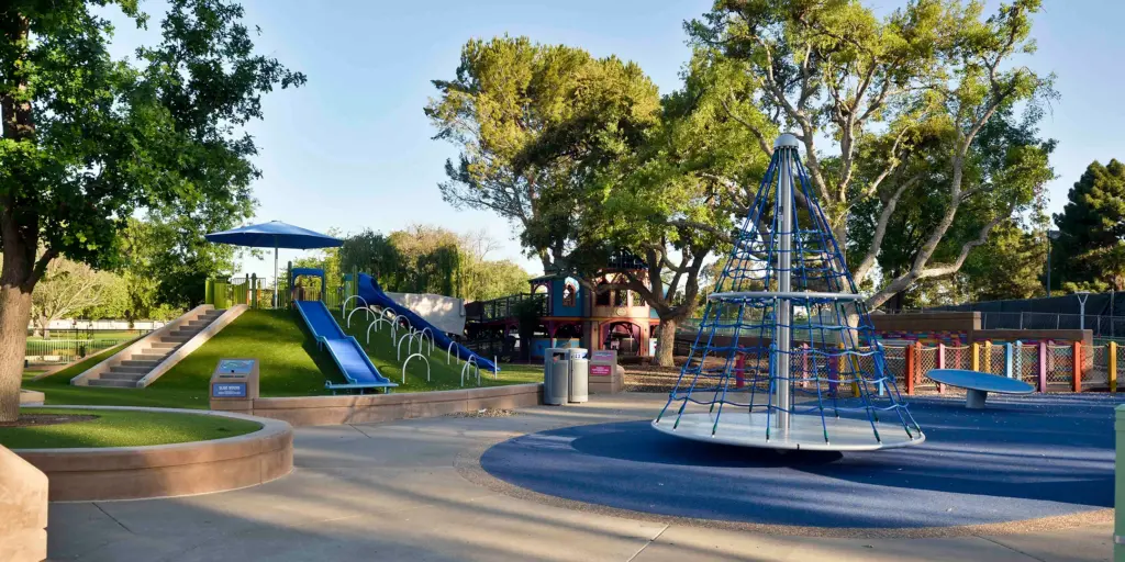 Playground with blue spinning equipment in the foreground, and a grassy hill with blue slides and a colorful playhouse in the background.