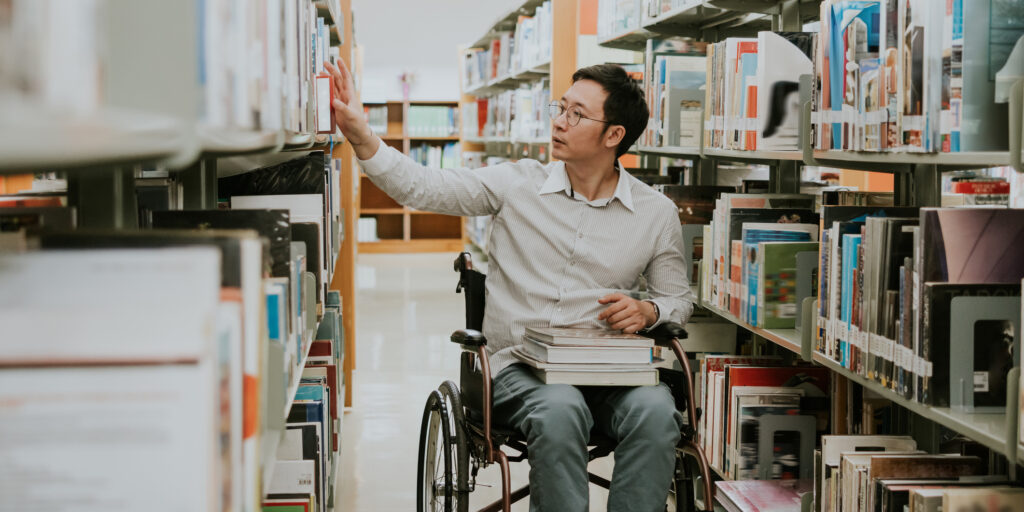 Asian man as a masters degree student with his wheelchair, looking and finding some books in university library for his work project.