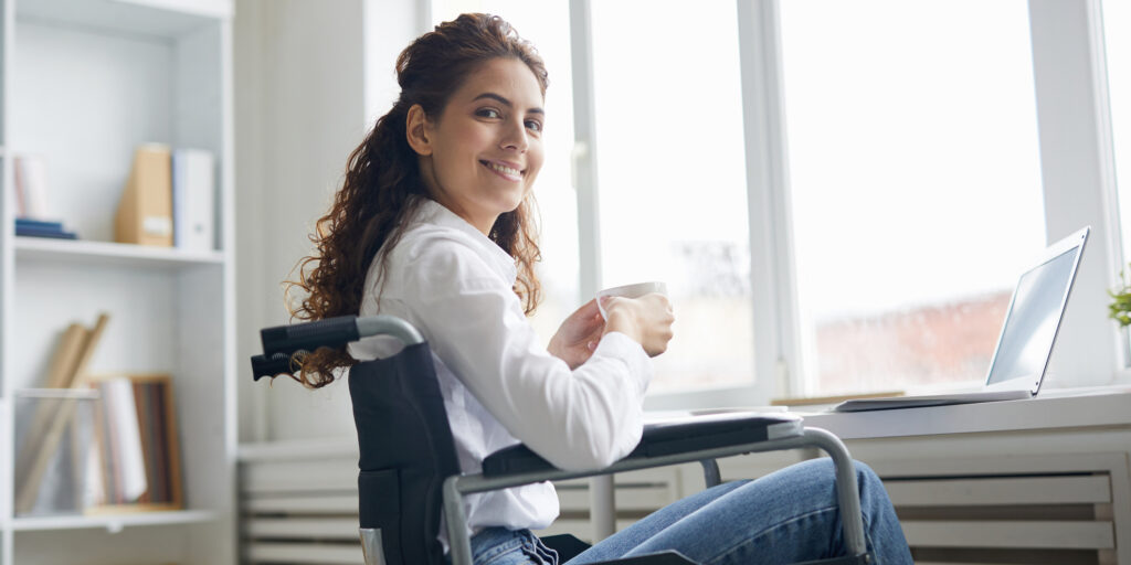 Young smiling manager with cup of tea looking at you while sitting by office window