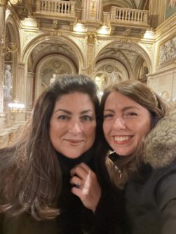 Two woman smile inside an opera house with detailed arches and statues