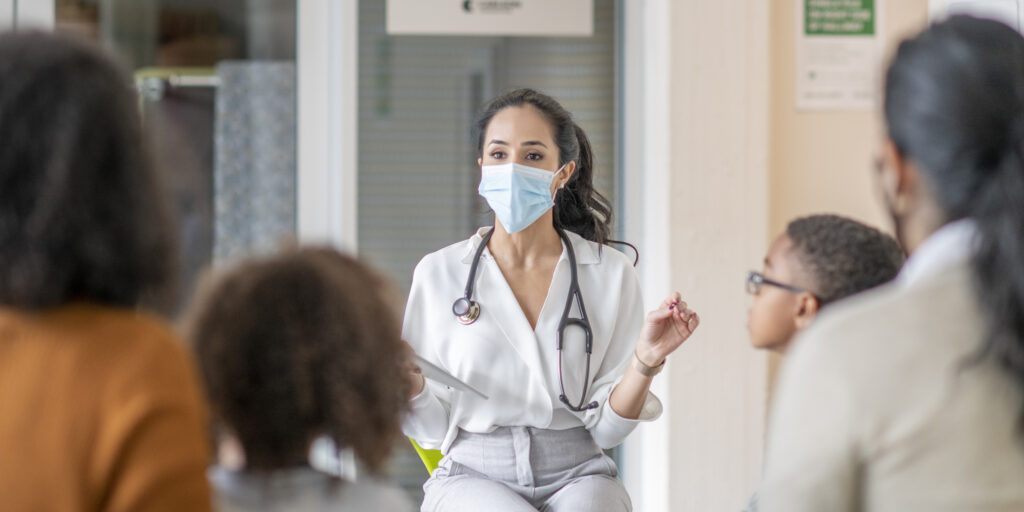 A doctor wearing a mask sits at the front of a room and addresses a group of women and children.