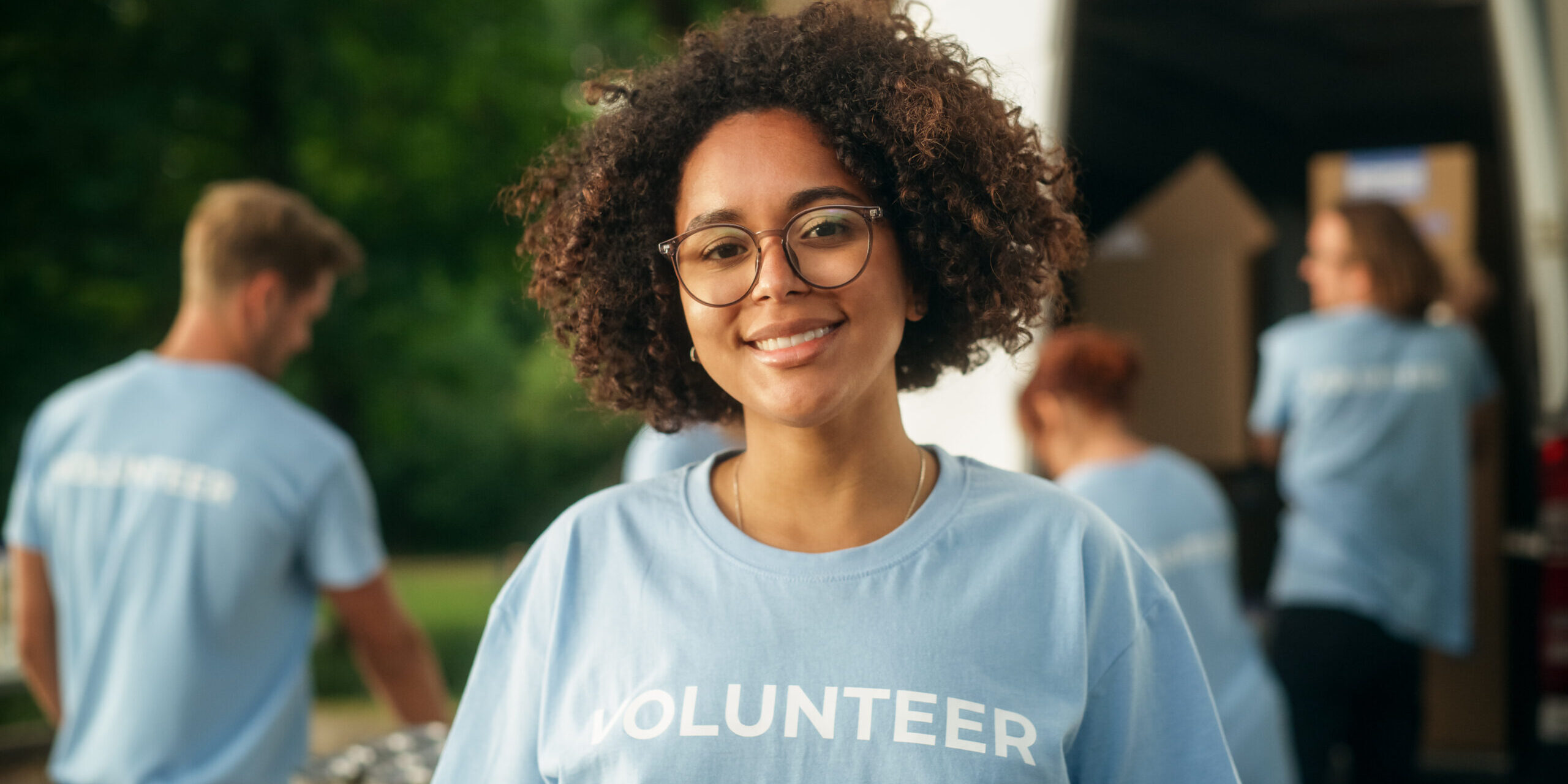 Young Adult Multiethnic Latina with Afro Hair, Wearing Glasses, Smiling, Wearing a shirt that says volunteer and Posing for Camera.