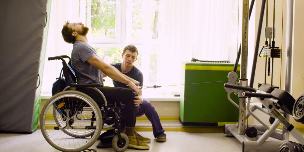 Young disabled man in wheelchair doing strength exercises for hands in the rehabilitation clinic. Doctor physiotherapist helping him