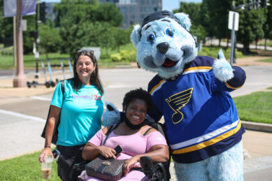 A woman in a wheelchair poses with another woman and a mascot at FestAbility