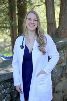A woman with long blonde hair smiles outside in a white medical coat with a stethoscope around her neck