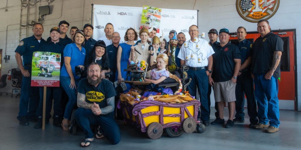 A young girl with a long blonde Rapunzel braid smiles in a wheelchair that has been modified to look like Rapunzel's tower is surrounded by friends, family, and fire fighters