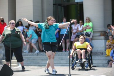 Three young women, two standing and one in a wheelchair, perform on stage with attendees watching in the background