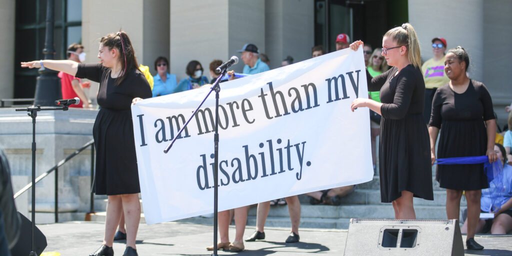 Dancers in black dresses hold a sign on stage that reads: I am more than my disability