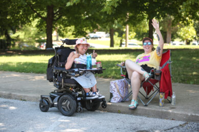 A woman in a powerwheelchair and a woman in a lawnchair sit outside and wave and give the peace sign to the camera man